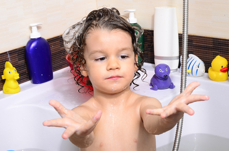Beautiful toddler taking a bath in a bathtub with bubbles. Cute kid washing his hair with shampoo in the shower and splashing water everywhere. Boy playing with toys in the tubの写真素材