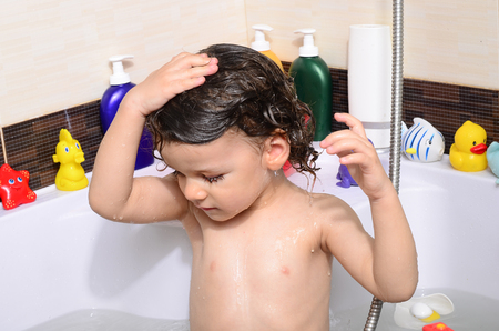 Beautiful toddler taking a bath in a bathtub with bubbles. Cute kid washing his hair with shampoo in the shower and splashing water everywhere. Boy playing with toys in the tubの写真素材
