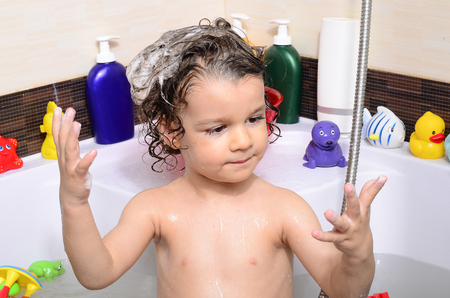 Beautiful toddler taking a bath in a bathtub with bubbles. Cute kid washing his hair with shampoo in the shower and splashing water everywhere. Boy playing with toys in the tubの写真素材