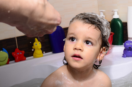 Beautiful toddler taking a bath in a bathtub with bubbles. Cute kid washing his hair with shampoo in the shower and splashing water everywhere. Boy playing with toys in the tubの写真素材