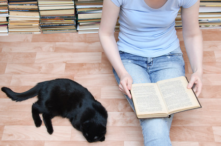 A girl sits on the floor and reads a book next to a black cat lying. Close-upの写真素材