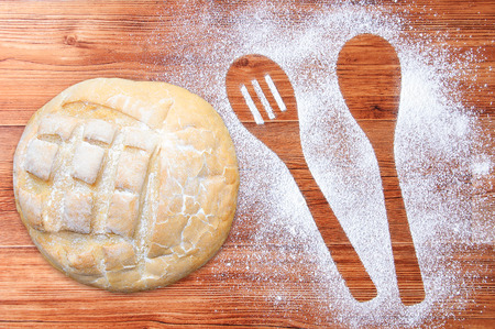 Wooden brown table dusted with flour and a loaf of wheat bread. Silhouette two spoons of flour.