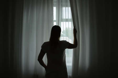 Caucasian woman at the window in the semi-darkness in the morning opens the curtains.の写真素材