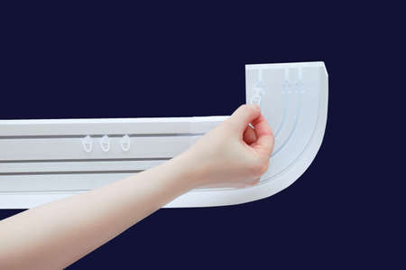 A Caucasian woman hand puts plastic hooks on a white curtain rod. Dark blue background.
