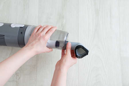 The hand of a caucasian woman puts on a small nozzle on a gray vacuum cleaner on a white wooden background.の写真素材