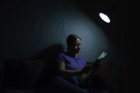 Caucasian young woman reading a book in the dark by the light of a lamp.