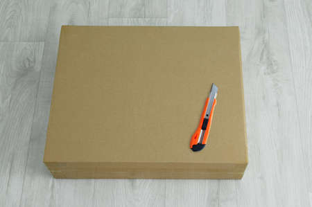 Brown cardboard box and a utility knife on the white wooden floor of the room.