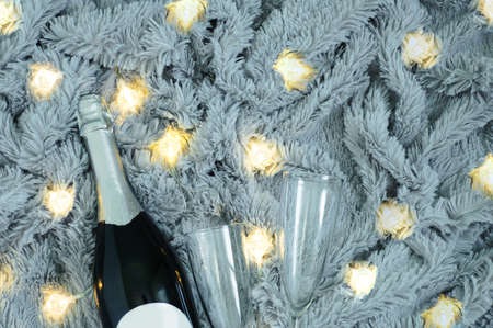 A bottle of champagne and two glasses against the background of a gray fur plaid with a luminous garland.の写真素材
