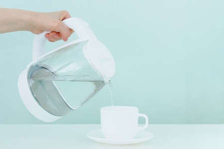 White glass teapot with hot water and drops of condensate. A hand pours hot water into a cup and saucer. green background.の素材 [FY310190133473]