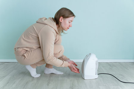 Caucasian woman warms herself by a fan heater while sitting on the floor of the room.の写真素材