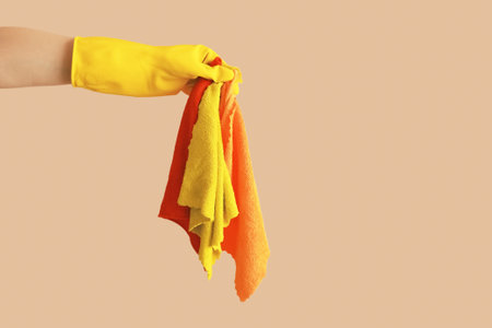 Female hand in yellow rubber protective glove holding three cleaning rags. Beige background mocha...