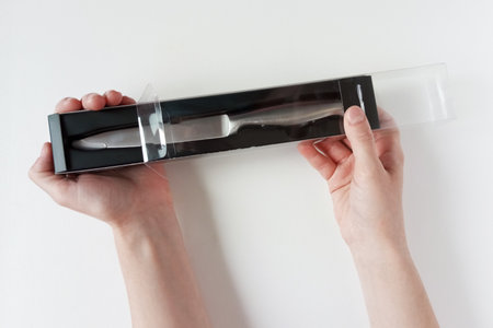 Caucasian woman takes out a new silver kitchen knife from the package. White background. Top view.