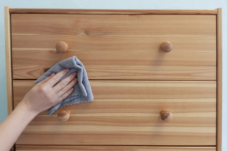 Caucasian woman's hand with gray cleaning cloth washing wooden chest of drawers. Dust removal, home...