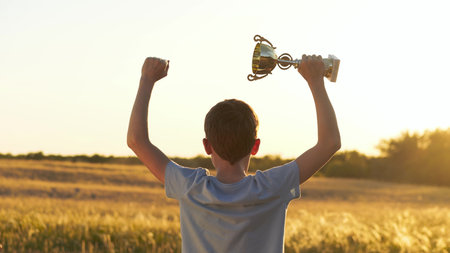 boy enjoys the open space of nature, raising trophy under warm sunlight. child feels free, joyful, and connected to the beauty of the outdoors. calm rural landscape enhances sense of peace and pride.の写真素材
