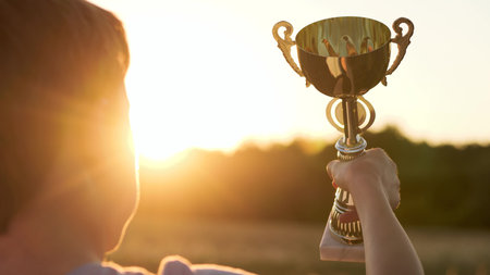 Small victor grips polished chalice against glowing horizon in vivid child victory. Bright silhouette lifts shining cup echoing kid triumph. Soft sunset beams infuse inspiring atmosphere child triumphの写真素材