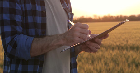 Close-up shot of agronomist hands holding clipboard and writing with pen in wheat field. Fieldwork documentation and crop research during golden hour. Modern farming and agricultural analysis.の写真素材