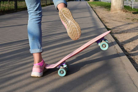 Close-up legs of girl skateboarder in blue jeans and pink sneakers, riding pink penny skate...