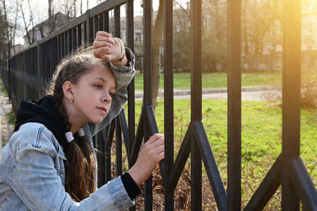 pretty, thoughtful girl in denim clothes is standing by a metal fence, leaning against it and...