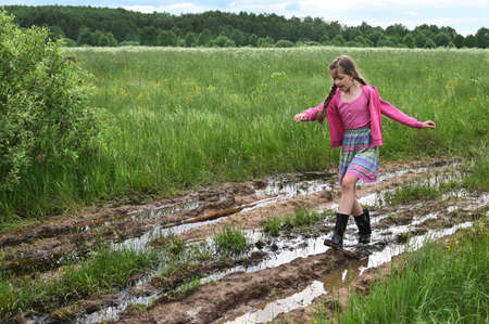 beautiful happy country girl in a pink dress and rubber boots runs and jumps in puddles. Muddy...