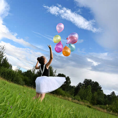 happy girl beautiful dress is holding colorful balloons in summer meadow against background of blue sky and clouds, having fun and playing. The concept of festive mood, birthday. Rear viewの写真素材
