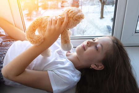 cute teenage girl is lying on the windowsill with a soft toy. The sun is shining outside the windowの写真素材