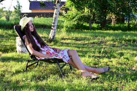 pretty girl with long wavy hair in hat is sitting on chaise longue against background of rural landscape. Summer outdoor recreationの写真素材