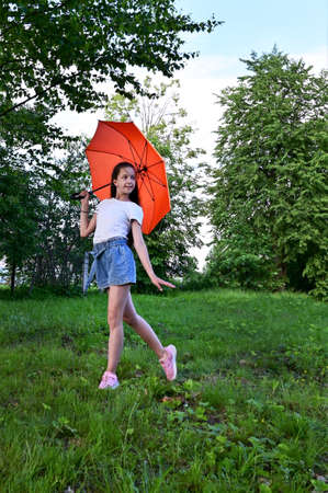 beautiful happy young girl with an orange umbrella is having fun on sunny summer day outdoors in park, dressed in white T-shirt and denim shorts. Travel, recreation, healthy lifestyleの写真素材