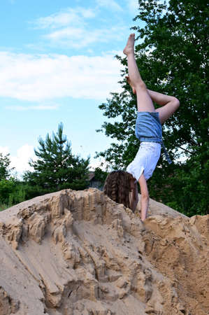 slender girl does handstand in open air against background of blue sky. Female athlete in white T-shirt and denim shorts performs acrobatic exercises. Copy spaceの写真素材
