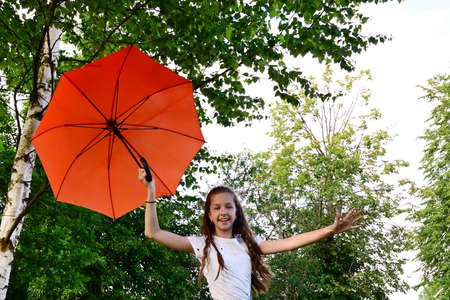 beautiful happy young girl in white T-shirt holds an orange umbrella, having fun on sunny summer day in park against the background of green foliage. Travel, recreation, healthy lifestyle. Copy spaceの写真素材