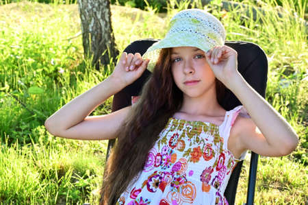 pretty girl with long wavy hair in hat is sitting on chaise longue against background of rural landscape. Summer outdoor recreationの写真素材