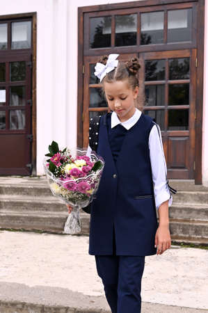 beautiful schoolgirl girl with white bow in her hair and beautiful school uniform with bouquet on the school porch. First day of school. Back to school. Knowledge Dayの写真素材