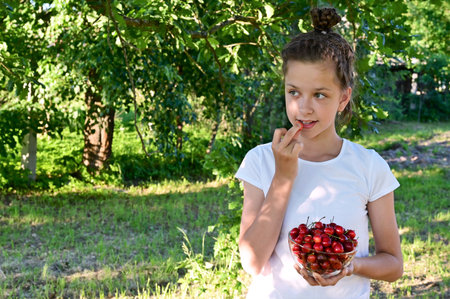 in summer garden young cute girl white T-shirt holds glass vase with ripe cherries and brings one berry to her mouth. Concept of gardening and harvesting. Delicious and healthy food. Copy spaceの写真素材