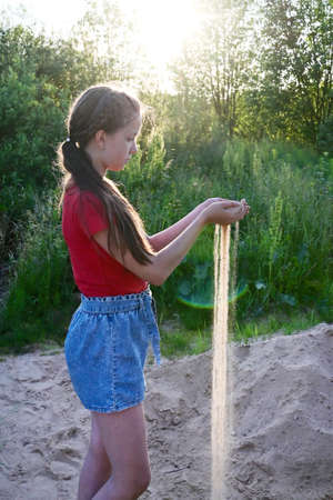 young girl in red T-shirt and denim shorts holds sand in her palms. The sand sifts through my fingers in the rays of setting sunの写真素材