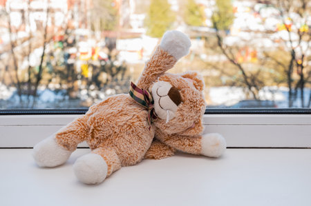 small plush toy kitten with a bow sits on a white windowsill and waves its paw. The sun is shining outside the window. Good morning wishesの写真素材