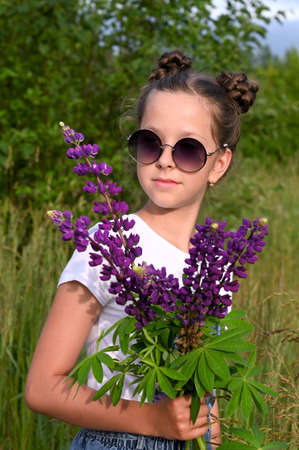 cute young girl wearing sunglasses holds a bouquet of purple lupin flowers near her face. Concept of nature, floristry and summer recreation. Close-up portraitの写真素材