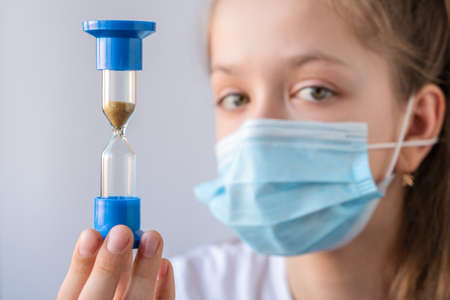 young girl in a medical mask holds an hourglass on a white background. The concept of time and its...