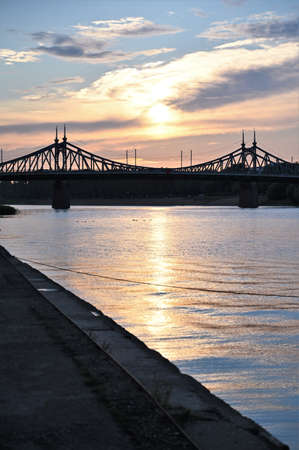 Urban landscape and a beautiful sunset in the evening on the river embankment with a bridge. Vertical photo. Copy spaceの写真素材