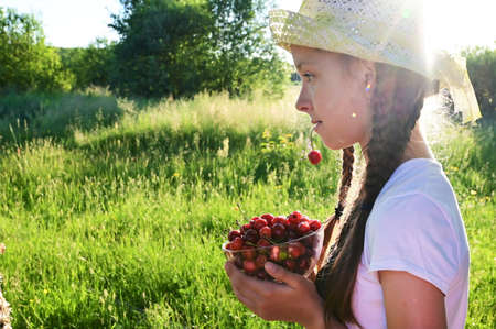 in summer garden young sweet girl in white T-shirt and hat holds ripe cherry berry in her mouth. Delicious and healthy foodの写真素材