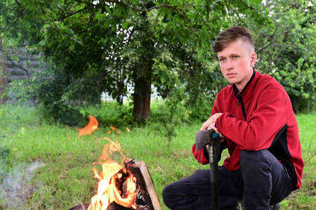 young serious man with an ax by fire in forest. Time for rest. Take a break. The path to nature. Copy spaceの写真素材