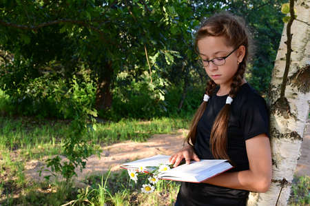 cute teenage girl with glasses is standing by tree in summer park and reading book. Ð¡oncept of training and education. Back to schoolの写真素材