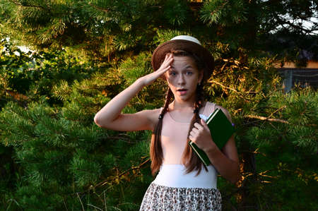An astonished beautiful teenage girl in hat and summer dress on background of green park with book...