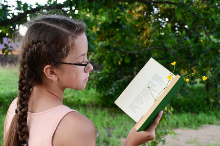 cute teenage girl with long braids and glasses is standing against background of green park and...