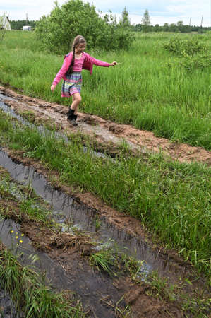 beautiful happy country girl in a pink dress and rubber boots runs and jumps in puddles. Muddy country road after rainの写真素材