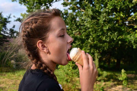 cute girl eats ice cream in waffle cone on background of green forest. Problem of hot climate and global warmingの写真素材