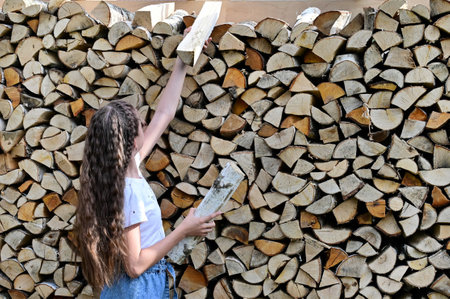 girl with long hair in white T-shirt and denim shorts near pile of firewood. Concept of harvesting...