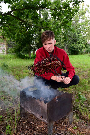 young man roasts meat, barbecue on coals on grill in nature. He's outside. Barbecue is located on green grass. There are trees in background. Concept of spending free time, joint barbecue, summer seasonの写真素材