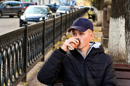 young man in jacket and baseball cap is sitting on bench and holding paper cup with drink in his hand. Guy, student, is drinking coffee against the background of autumn urban landscape. Rest, break. Takeaway coffeeの写真素材