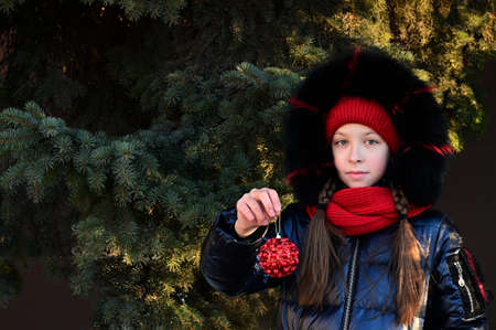 girl at natural Christmas tree with a Christmas toy in her hands in winter. Preparing for New Year,...