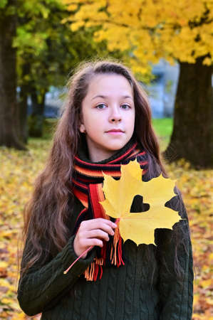 pretty girl in sweater and scarf holds yellow maple leaf with heart carved on it. Teenage girl walks in autumn park and enjoys nature. Vertical photoの写真素材