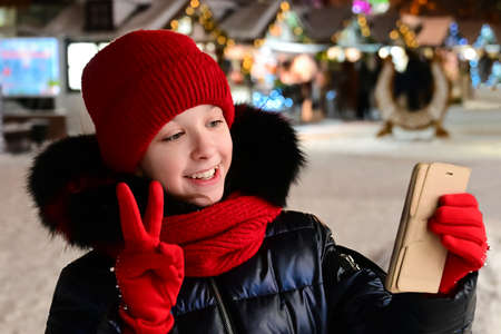 pretty girl in red hat, scarf and gloves is talking phone via video link and showing victory sign "V" with her fingers against background winter New Year decorated street. Wishes for Christmasの写真素材